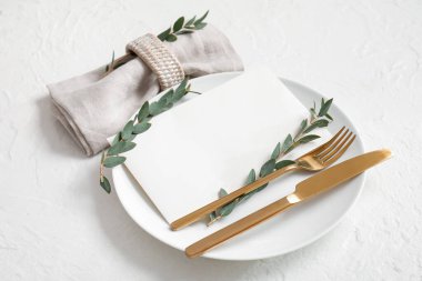 Boş davetiye kartı, çatal bıçak takımı ve beyaz masa üzerinde okaliptüs dallarıyla temiz tabak.