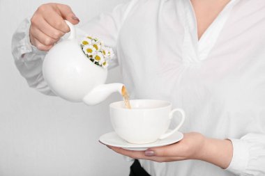 Güzel genç bir kadın çaydanlıktan beyaz duvarın yanındaki fincana papatya çayı dolduruyor.