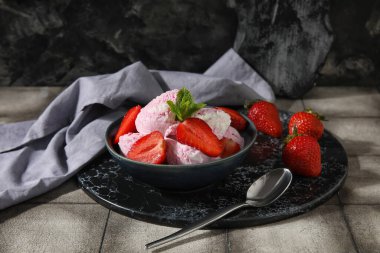 Bir kase çilekli dondurma, nane ve kaşık ile siyah grunge duvarın yanındaki gri fayans masasında.