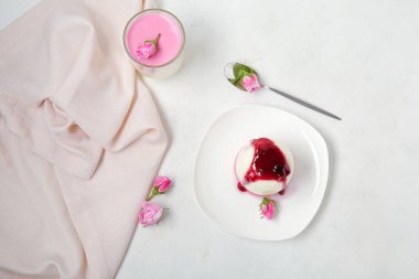 Beyaz masa üzerinde pembe gül çiçekleri olan bir tabak ve bir bardak panna cotta.