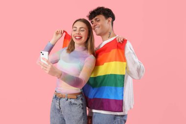 LGBT bayrağı taşıyan genç bir çift pembe arka planda selfie çekiyor.