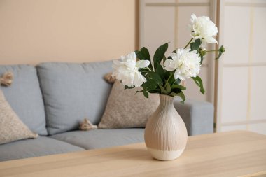 Kahve masası, kanepe ve bej duvarın yanında tuvalet ekranı olan beyaz şakayık vazosu.