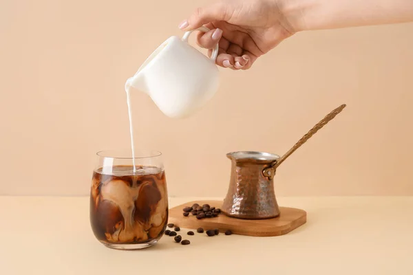 女人用米色背景的冰咖啡把牛奶从罐子倒进杯子里 — 图库照片