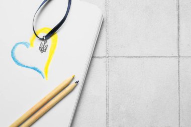 Beyaz karo arka planda Ukrayna arması, defter, çizilmiş kalp ve kalemlerle kurdele.