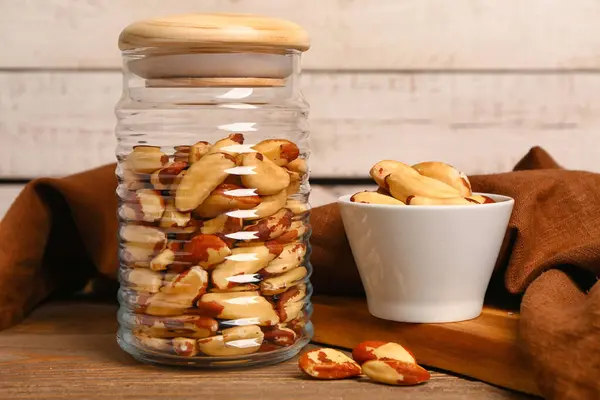 stock image Glass jar and bowl of tasty Brazil nuts on wooden table