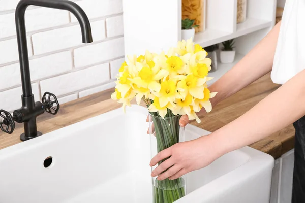 Woman taking vase with narcissus flowers from sink, closeup