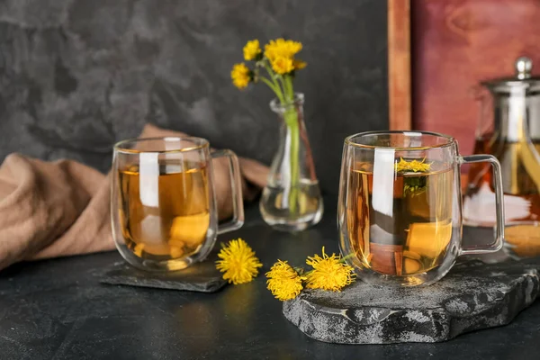 stock image Board with glass cups of healthy dandelion tea and teapot on black table