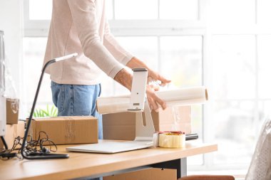 Genç adam ofisinde streç filmle karton kutu paketliyor.