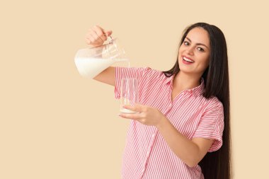 Güzel genç bir kadın bej arka planda bardağa süt döküyor.