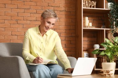 Evde dizüstü bilgisayar ve dizüstü bilgisayarla çalışan olgun bir kadın serbest yazar.