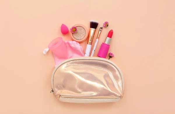Lady's cosmetic bag with makeup products, menstrual cup and sponge on pale orange background