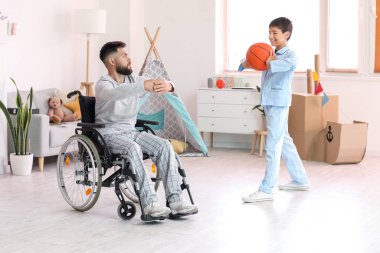 Küçük çocuk ve babası tekerlekli sandalyede evde top oynuyorlar.