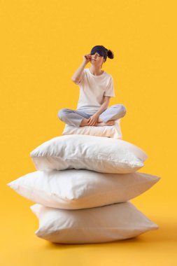 Young woman with sleeping mask and soft pillows sitting on yellow background