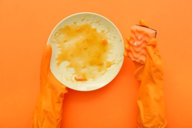 Lastik eldivenli kadın elleri turuncu arka planda kirli tabakları süngerle yıkıyor.