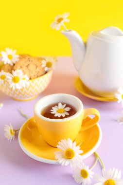 Sarı duvarın yanındaki pembe masada doğal papatya çayı, kurabiye ve çiçeklerle çaydanlık.