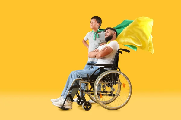 男の子と彼の父で車椅子スーパーヒーローの帽子をかぶって黄色の背景 — ストック写真