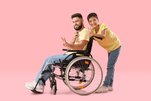 ピンクの背景に勝利のジェスチャーを示す車椅子の男の子と彼の父親 — ストック写真