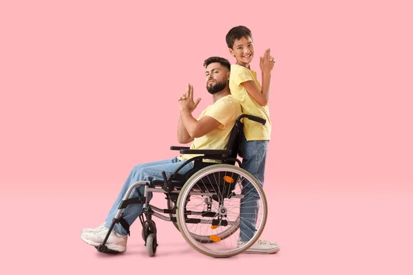 ピンクの背景に銃のジェスチャーを示す車椅子の男の子と彼の父親 — ストック写真
