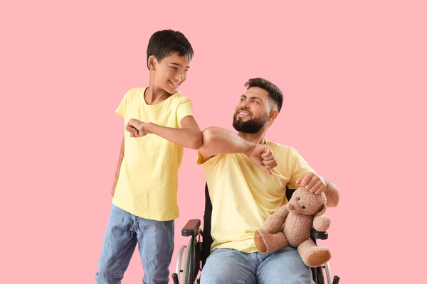 小さな男の子と彼の父で車椅子におもちゃバンピング肘でピンクの背景 — ストック写真