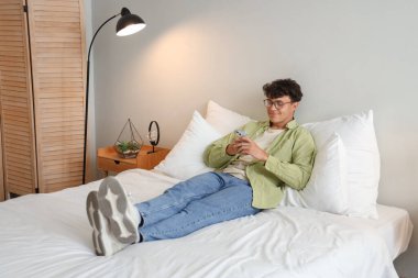 Yatak odasında cep telefonu kullanan gözlüklü genç adam.