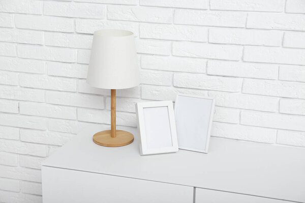 White chest of drawers with lamp and blank pictures near light brick wall in room, closeup