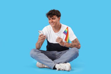 LGBT bayrağı taşıyan genç adam mavi arka planda cep telefonu kullanıyor