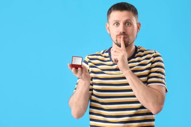 Nişan yüzüklü yakışıklı adam mavi arka planda sessizlik işareti yapıyor.