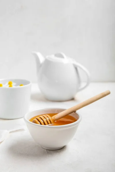 stock image Bowl of honey, teapot and cup with chamomile flowers on light background