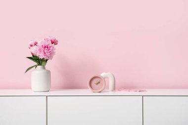 Pembe duvarın yanında şakayık çiçekleri ve çalar saati olan şık beyaz çekmeceler, yakın plan.