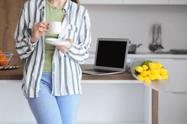 Masanın yanında modern laptop ve sarı lale çiçekleriyle kahve içen bir kadın.