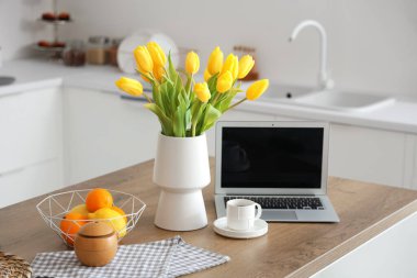 Sarı lale çiçekli vazo, modern laptop, meyveler ve açık mutfak masasında bir fincan kahve.