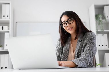 Gözlüklü güzel Asyalı iş kadını ofiste dizüstü bilgisayarla çalışıyor.