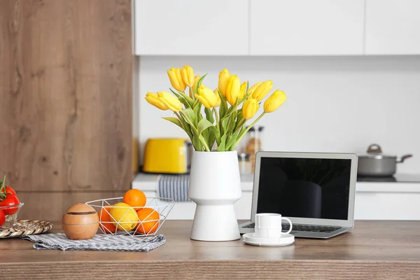 黄色のチューリップの花の花瓶 現代のラップトップ 果物や軽いキッチンで木製のテーブルの上にコーヒーのカップ — ストック写真