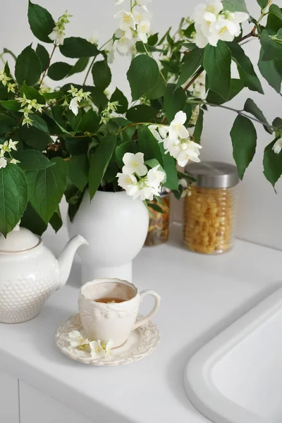 Vase with blooming jasmine flowers, teapot and cup of tea on white kitchen counter