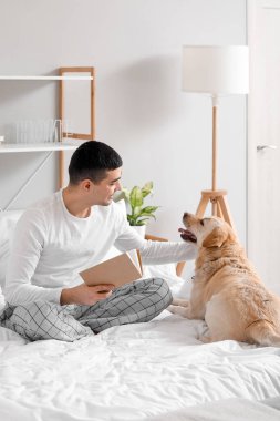 Kitabı olan genç bir adam ve yatak odasında oturan sevimli Labrador köpeği.
