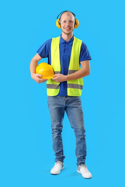 Male worker in vest, with hardhat and protective headphones on blue background