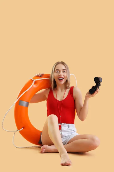 Female lifeguard with binoculars and ring buoy sitting on beige background