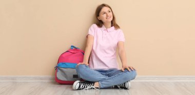 Okul çantalı genç kız bej rengi duvarın yanında oturuyor.