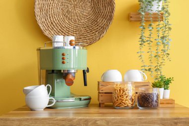 Modern kahve makinesi, fincanlar ve atıştırmalıklar sarı duvarın yanındaki masada.