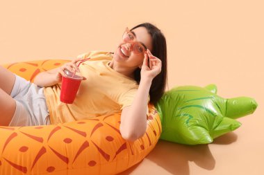 Bir bardak soda ve bej arka planda duran şişirilebilir yüzüklü genç kadın.