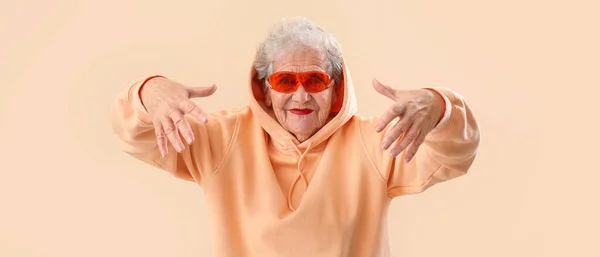 Stilvolle Seniorin Mit Sonnenbrille Und Kapuzenpulli Auf Beigem Hintergrund — Stockfoto
