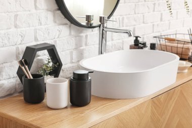 Lavabo kasesi ve banyo aksesuarları beyaz tuğla duvarın yanındaki ahşap dolapta.