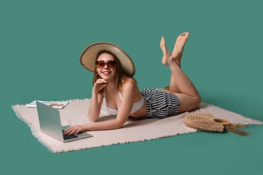 Plaj aksesuarları olan genç bir kadın yeşil arka planda dizüstü bilgisayar kullanıyor.