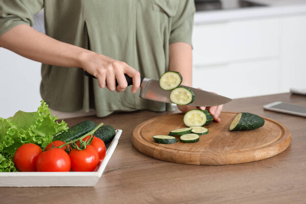 Woman cutting cucumber for salad on wooden table with vegetables