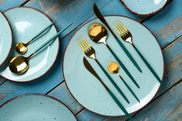青い木製のテーブルの上に黄金のカトラリーのセットときれいなプレート — ストック写真