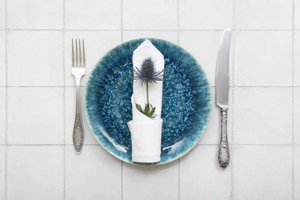 白いタイル張りのテーブルの上にきれいなプレート エルニジウムの花とカトラリーと美しいテーブルの設定 — ストック写真