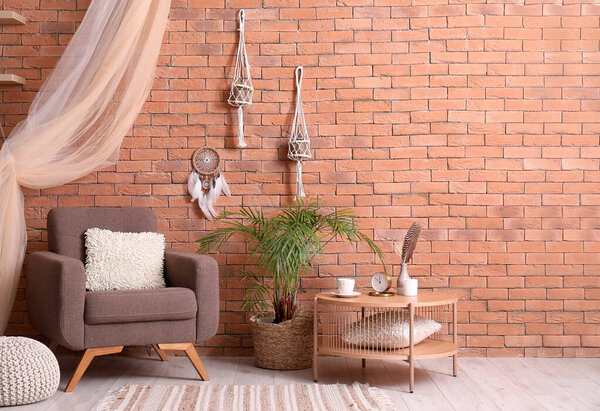 Interior of modern living room with armchair, table, plant and dream catcher hanging on brick wall