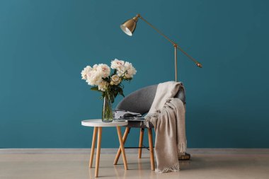 Kahve sehpası, sandalye ve yer lambası olan beyaz şakayık vazosu mavi duvarın yanında.