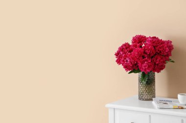 Bej duvarın yanındaki tuvalet masasında bir fincan kahve ve gazete bulunan kırmızı şakayık vazosu.