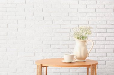 Güzel çiçekli vazo ve beyaz tuğla duvarın yanındaki ahşap sehpanın üzerinde fincan.